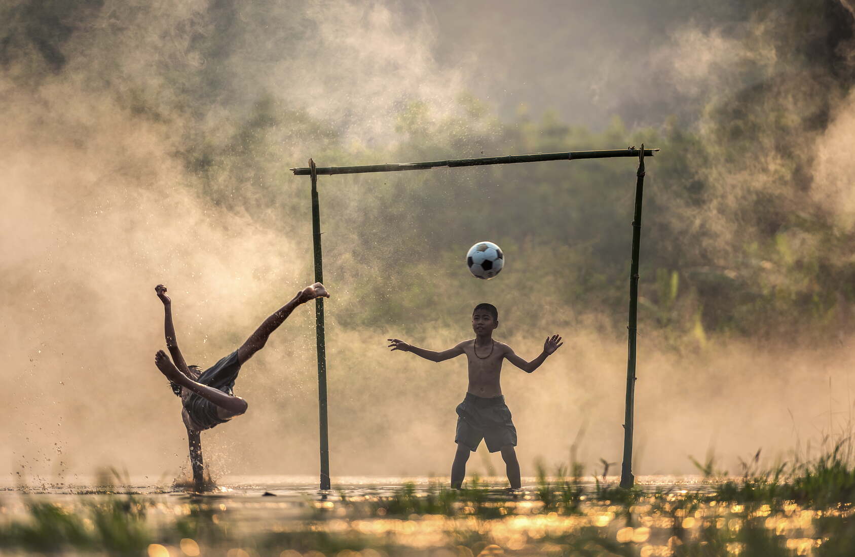 Imagen de chico descalzo y en el agua pateando de tijera una pelota a otro que esta atajando